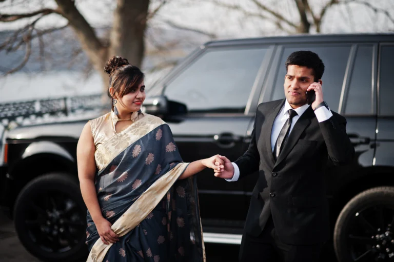 Elegant and fashionable indian friends couple of woman in saree and man in suit posed against rich black suv car. Guy speaking on mobile phone.
